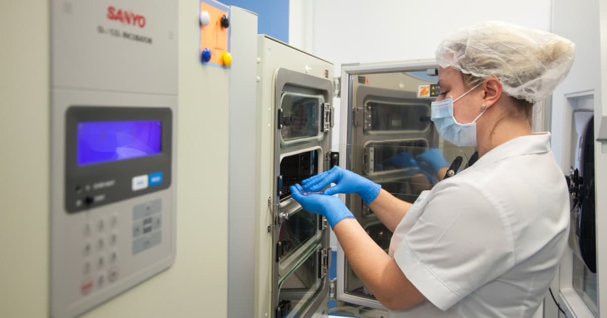 An embryologist in a hairnet and gloves placing a culture dish into an incubator in a fertility clinic laboratory